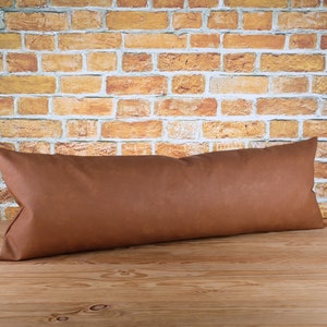 May include: A long, rectangular brown cushion with a soft, textured appearance. The cushion is placed on a wooden surface, with a brick wall in the background. The cushion's shape suggests it is designed for lumbar support or as a decorative element.