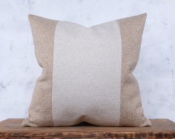 Op de afbeelding: Vierkant decoratief kussen met neutrale kleuren. Het heeft een centraal paneel van lichtbeige stof, geflankeerd door panelen van donkerder beige stof. Het kussen ligt op een houten oppervlak, wat een eenvoudige uitstraling geeft.