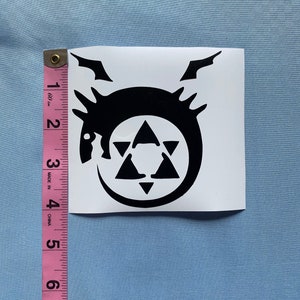 May include: A black vinyl sticker featuring a circular design with a dragon-like creature, a six-pointed star, and a crescent moon. The sticker is approximately 3 inches (7.6 cm) in height and width.