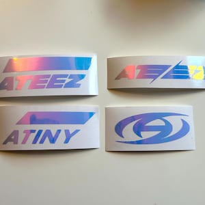 May include: Four rectangular stickers with iridescent holographic designs. Two stickers display the word "ATEEZ" in a stylized font, while another says "ATINY". The fourth sticker features a stylized design. All stickers have a white background.