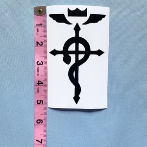 May include: A black and white sticker featuring a stylized alchemical symbol. The sticker is approximately 5 inches tall and includes a crown, wings, a cross, and a snake. The sticker is on a white background.