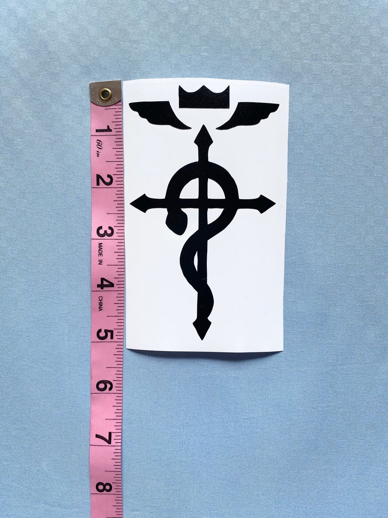May include: A black and white sticker featuring a stylized alchemical symbol. The sticker is approximately 5 inches tall and includes a crown, wings, a cross, and a snake. The sticker is on a white background.