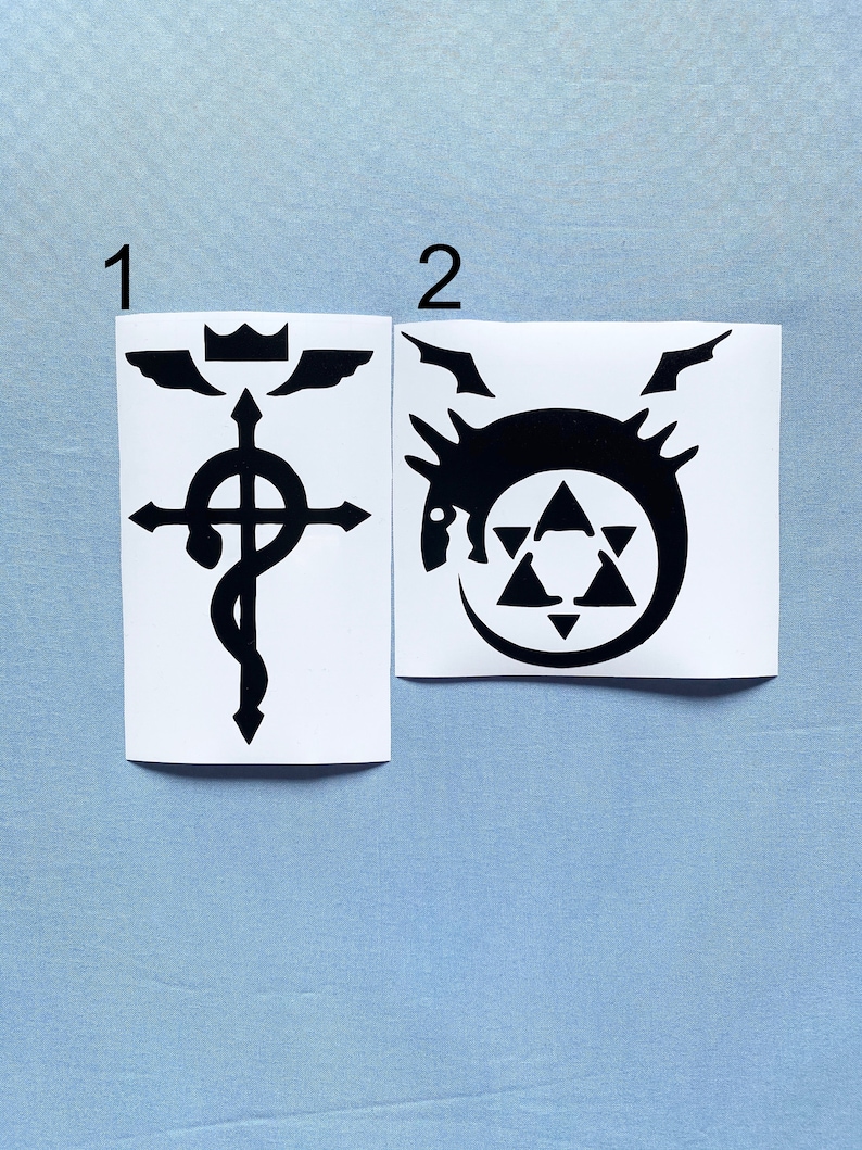 May include: Two black and white vinyl stickers. The first features a cross with a serpent wrapped around it, topped with a crown and wings. The second sticker displays a dragon-like creature within a circle, with a star-like shape inside.