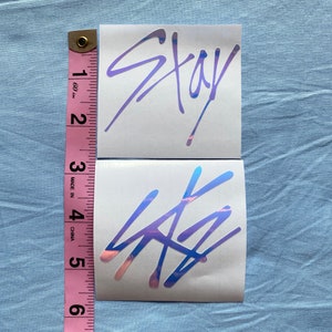 Peut inclure: Deux autocollants holographiques avec le mot "Stay" et un logo dans une couleur iris&eacute;e violette et bleue. Les autocollants sont sur un fond blanc.