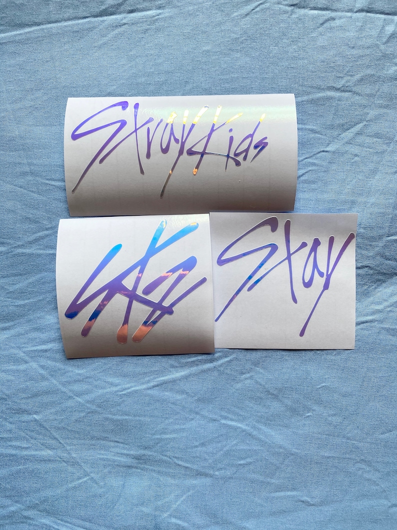 Peut inclure: Trois autocollants blancs avec des lettres holographiques violettes et dor&eacute;es. L'autocollant du haut dit "Stray Kids". L'autocollant du milieu a un logo stylis&eacute;. L'autocollant du bas dit "Stay".
