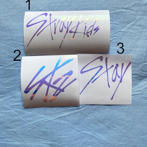 Peut inclure: Trois autocollants blancs avec des lettres holographiques violettes et argent&eacute;es. Le premier autocollant dit "Stray Kids". Le deuxi&egrave;me autocollant a un logo stylis&eacute;. Le troisi&egrave;me autocollant dit "Stay".
