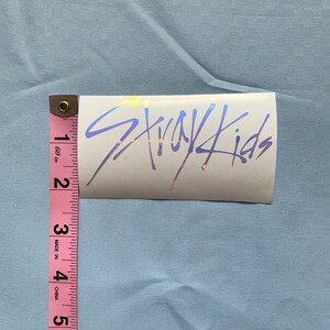 Peut inclure: Un autocollant blanc avec un logo holographique bleu et jaune qui dit "Stray Kids". L'autocollant mesure environ 5 cm de large et 2,5 cm de haut.