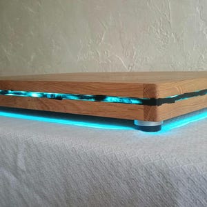 Op de afbeelding: Een rechthoekige houten serveerplank met een horizontale spleet gevuld met lichtgevende cyaan hars. De plank staat op vier kleine, ronde zilverkleurige voetjes die een fel cyaan licht werpen op het oppervlak onder het object.