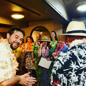 May include: A group of people wearing colorful Hawaiian shirts and leis, smiling and laughing. Some are holding white cards.