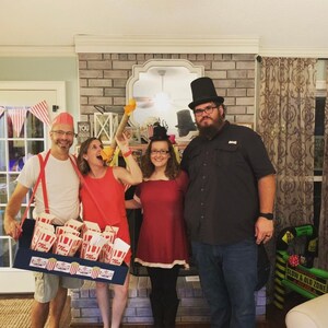 May include: Four people dressed in costumes for a carnival or circus themed party. A person wearing a red and white striped shirt and suspenders holds a tray of popcorn. A person wearing a red dress and a top hat holds a red and white striped stick. A person wearing a red dress and a top hat holds a red and white striped stick. A person wearing a black shirt and a top hat stands behind the group.