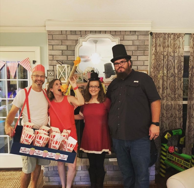 May include: Four people dressed in costumes for a carnival or circus themed party. A person wearing a red and white striped shirt and suspenders holds a tray of popcorn. A person wearing a red dress and a top hat holds a red and white striped stick. A person wearing a red dress and a top hat holds a red and white striped stick. A person wearing a black shirt and a top hat stands behind the group.