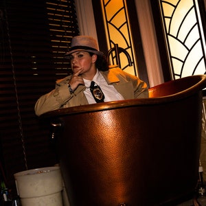 May include: A person wearing a fedora and a trench coat sits in a copper bathtub. The bathtub is in a room with a window behind the person.