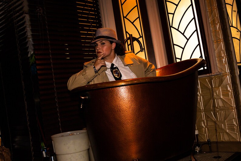 May include: A person wearing a fedora and a trench coat sits in a copper bathtub. The bathtub is in a room with a window behind the person.