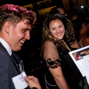 May include: Two people are laughing at a party. The woman is wearing a black dress with sequins and a pearl necklace. The man is wearing a dark suit and a name tag that says "David".