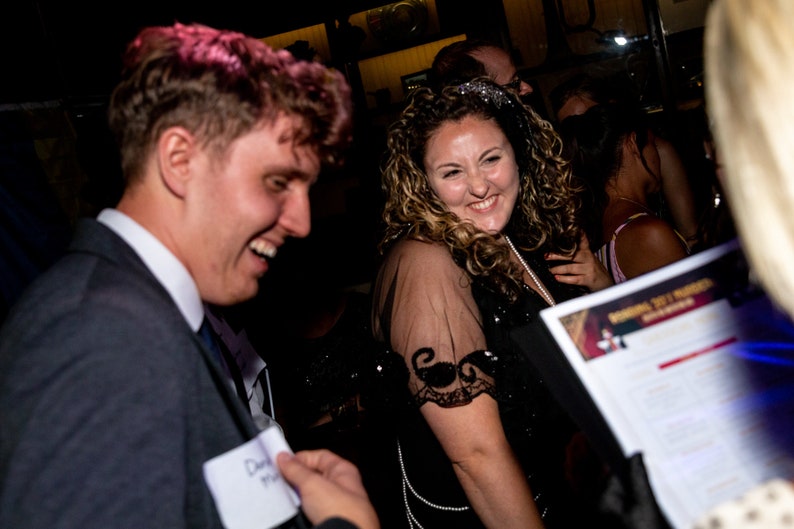 May include: Two people are laughing at a party. The woman is wearing a black dress with sequins and a pearl necklace. The man is wearing a dark suit and a name tag that says "David".
