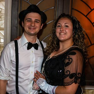 May include: A couple dressed in 1920s attire. The man is wearing a white shirt, a black bow tie, a black fedora, and suspenders. The woman is wearing a black beaded dress with a lace overlay and a white headband. They are standing in front of a window with a stained glass design.