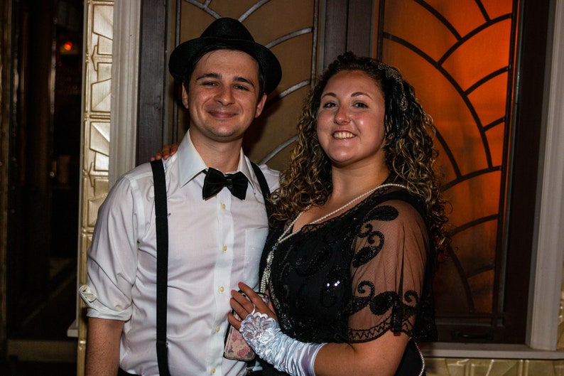 May include: A couple dressed in 1920s attire. The man is wearing a white shirt, a black bow tie, a black fedora, and suspenders. The woman is wearing a black beaded dress with a lace overlay and a white headband. They are standing in front of a window with a stained glass design.
