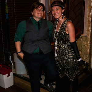May include: Two people dressed in 1920s attire. A person wearing a green shirt, a gray vest, and black pants. A person wearing a green and gold beaded dress with a pearl necklace and black gloves.