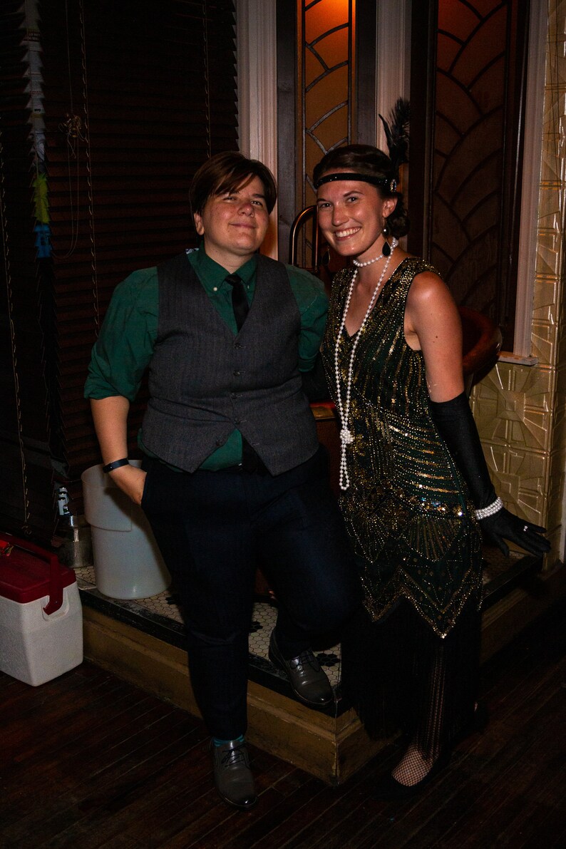 May include: Two people dressed in 1920s attire. A person wearing a green shirt, a gray vest, and black pants. A person wearing a green and gold beaded dress with a pearl necklace and black gloves.
