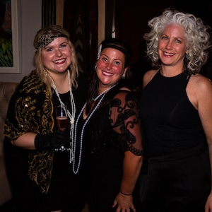 May include: Three women wearing black dresses and headbands pose for a photo. The woman on the left is holding a glass of champagne. The woman in the middle is wearing a pearl necklace. The woman on the right has gray hair.