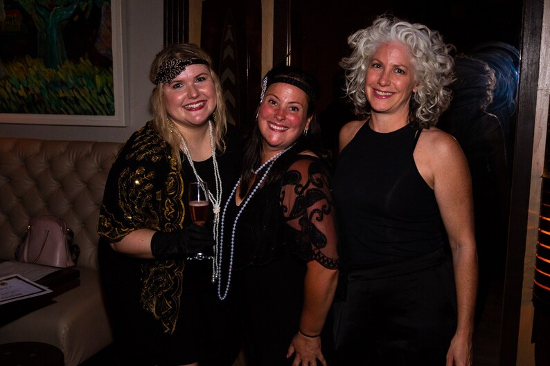 May include: Three women wearing black dresses and headbands pose for a photo. The woman on the left is holding a glass of champagne. The woman in the middle is wearing a pearl necklace. The woman on the right has gray hair.
