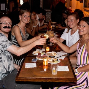 May include: A group of friends toasting with cocktails at a table with a spread of appetizers, including cheese, crackers, and olives. The table has a copper-colored surface and is set with napkins and glasses.