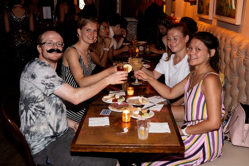 May include: A group of friends toasting with cocktails at a table with a spread of appetizers, including cheese, crackers, and olives. The table has a copper-colored surface and is set with napkins and glasses.