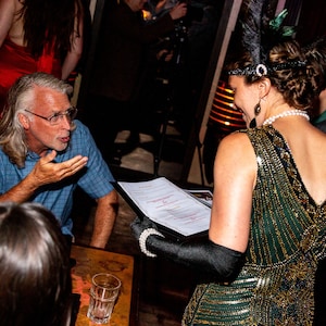 May include: A man with grey hair and glasses is talking to a woman wearing a green sequined dress and a feather headband. The woman is holding a menu.