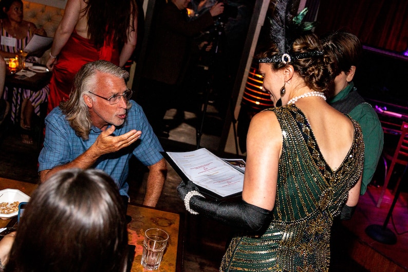 May include: A man with grey hair and glasses is talking to a woman wearing a green sequined dress and a feather headband. The woman is holding a menu.