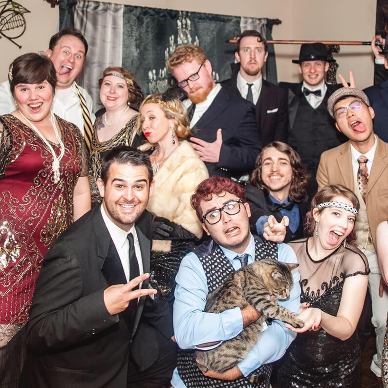 May include: A group of people dressed in 1920s attire, including a man holding a cat, a woman wearing a sequined dress, and a man wearing a tuxedo. The group is posing for a photo in a room decorated with a vintage aesthetic.