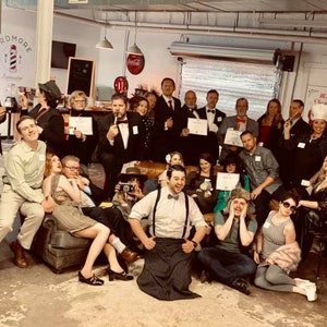 May include: A group of people dressed in various costumes and attire, some holding certificates, gathered in a room with a sign that reads "Ardmore Barbershop".