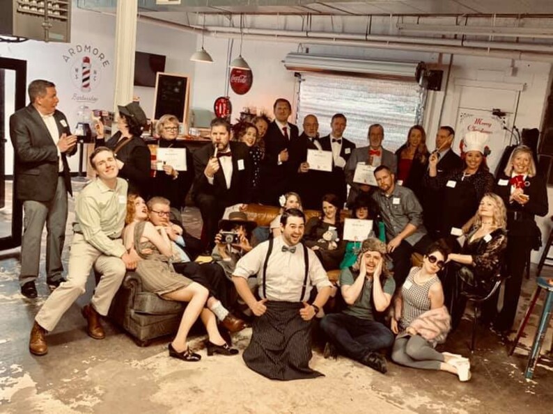 May include: A group of people dressed in various costumes and attire, some holding certificates, gathered in a room with a sign that reads "Ardmore Barbershop".
