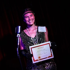 May include: A woman in a sequined green dress and a pearl necklace holds a certificate that reads "Broadway Certificate of Appreciation to [illegible] for [illegible]" with a red star in the corner. She is smiling and wearing a headband and black gloves. A microphone is in front of her.