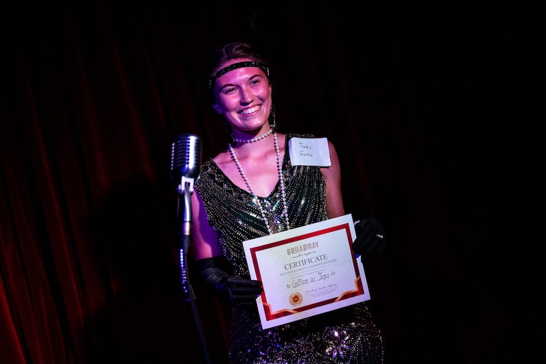 May include: A woman in a sequined green dress and a pearl necklace holds a certificate that reads "Broadway Certificate of Appreciation to [illegible] for [illegible]" with a red star in the corner. She is smiling and wearing a headband and black gloves. A microphone is in front of her.