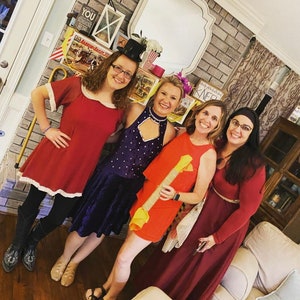May include: Four women dressed in costumes for a party. The woman on the left is wearing a red dress with white trim, the woman in the middle is wearing a purple dress with sequins, the woman on the right is wearing a red dress with a black belt, and the woman in the back is wearing a red dress with a white collar.