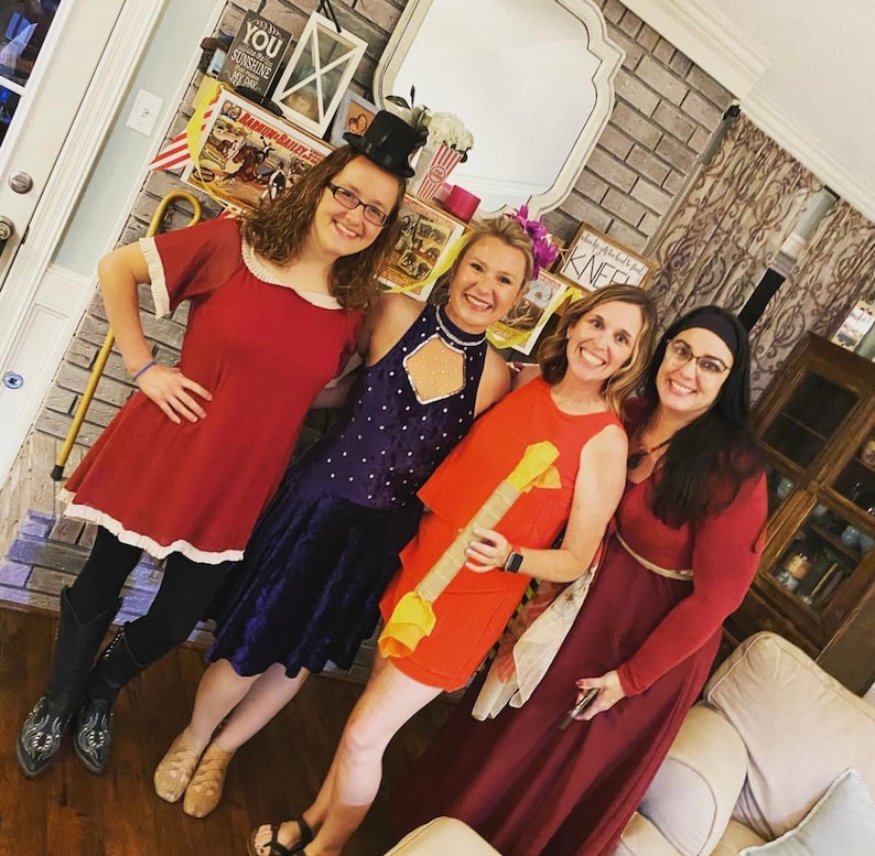 May include: Four women dressed in costumes for a party. The woman on the left is wearing a red dress with white trim, the woman in the middle is wearing a purple dress with sequins, the woman on the right is wearing a red dress with a black belt, and the woman in the back is wearing a red dress with a white collar.
