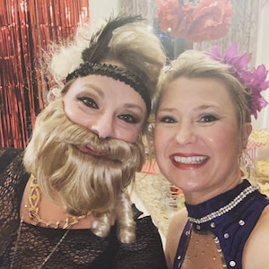 May include: Two women dressed in costumes for a party. One woman is wearing a black dress with a gold chain necklace and a blonde wig with a beard. The other woman is wearing a purple dress with rhinestones and a flower in her hair.