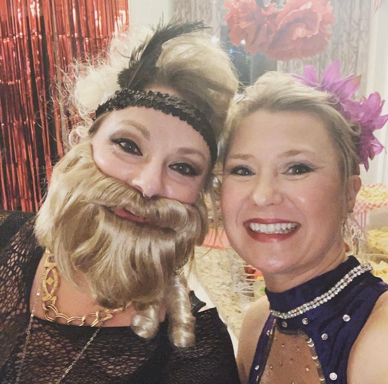 May include: Two women dressed in costumes for a party. One woman is wearing a black dress with a gold chain necklace and a blonde wig with a beard. The other woman is wearing a purple dress with rhinestones and a flower in her hair.