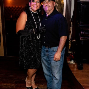 May include: A couple dressed in 1920s attire. The woman is wearing a black sequined dress with a pearl necklace and a feather headpiece. The man is wearing a black shirt, blue jeans, and a newsboy cap.