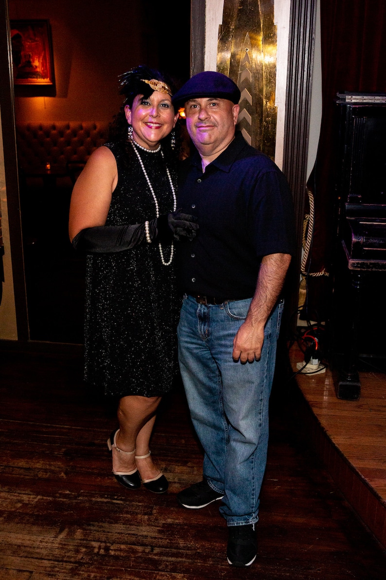 May include: A couple dressed in 1920s attire. The woman is wearing a black sequined dress with a pearl necklace and a feather headpiece. The man is wearing a black shirt, blue jeans, and a newsboy cap.