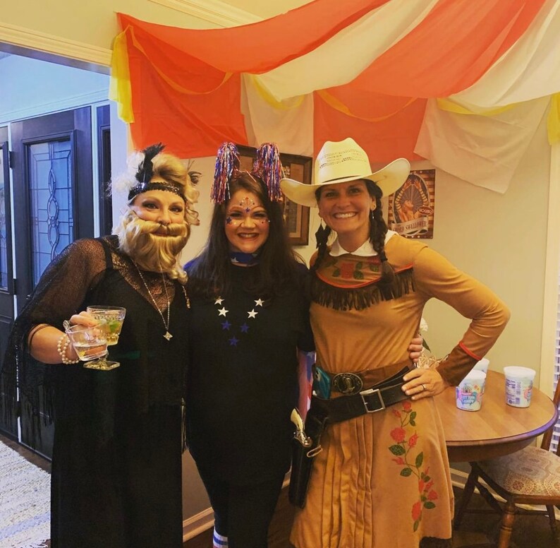 May include: Three women dressed in costumes for a party. One woman is wearing a black dress with a feather boa and a fake beard. Another woman is wearing a black shirt with stars and a blue headband. The third woman is wearing a brown cowgirl costume with a white cowboy hat.