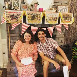 May include: A couple poses in front of a brick fireplace decorated with red and white striped bunting and vintage circus posters. The posters feature images of elephants, clowns, and other circus performers. The text on the posters reads "Barnum & Bailey" and "When the gifts are too hard to stand, kneel".