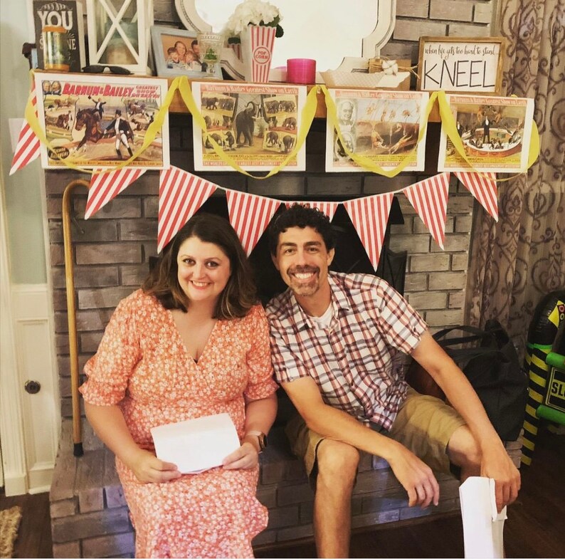May include: A couple poses in front of a brick fireplace decorated with red and white striped bunting and vintage circus posters. The posters feature images of elephants, clowns, and other circus performers. The text on the posters reads "Barnum & Bailey" and "When the gifts are too hard to stand, kneel".