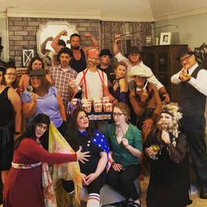 May include: A group of people dressed in costumes for a party. Some are wearing hats, others have facial hair, and one person is holding a box of popcorn. The group is standing in a living room with a couch and a coffee table.