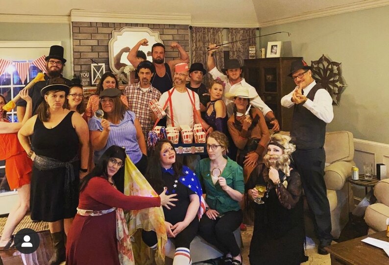 May include: A group of people dressed in costumes for a party. Some are wearing hats, others have facial hair, and one person is holding a box of popcorn. The group is standing in a living room with a couch and a coffee table.