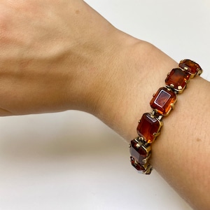 Op de afbeelding: Een vintage armband met grote, rechthoekige barnsteenkleurige stenen gezet in goudkleurig metaal. De armband wordt getoond op een pols, waarbij de warme tinten van de stenen en de gedetailleerde zetting worden benadrukt.