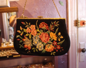 May include: Black handbag with a floral needlepoint design. The bag has a gold chain strap and clasp. The needlepoint design features colourful flowers in shades of pink, yellow, orange, and blue.