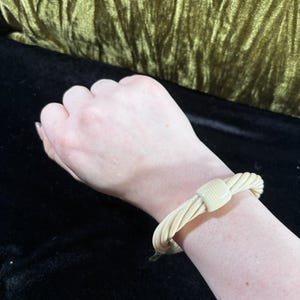 May include: A cream-coloured bracelet with a twisted rope design. The bracelet is secured with a rectangular clasp. The jewellery is worn on a wrist, with a black background and a gold-coloured fabric in the background.