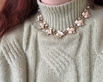 May include: A gold-toned necklace with white enamel leaf-shaped pendants and small clear rhinestones. The necklace is worn over a light green cable-knit sweater.