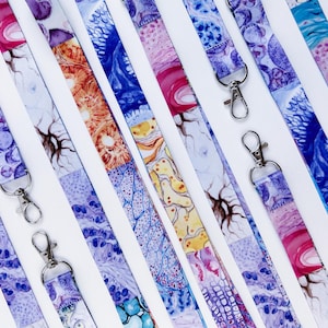 May include: A collection of lanyards with colorful abstract patterns. Each lanyard features a different design with shades of blue, purple, pink, and yellow. They have silver-colored metal clasps for attaching keys or ID badges.